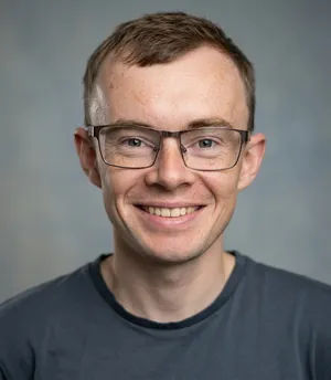 James Devine's headshot. A middle aged white man with short brown hair smiling at the camera. He is wearing a blue shirt and a red hoodie.