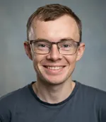 James Devine's headshot. A middle aged white man with short brown hair smiling at the camera. He is wearing a blue shirt and a red hoodie.